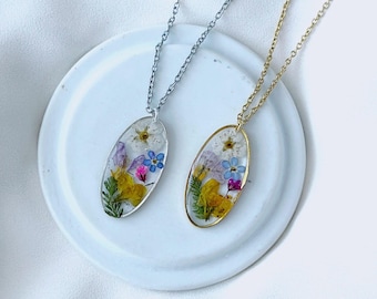 May include: Two oval pendant necklaces with dried flowers encased in clear resin. One necklace has a silver chain, the other a gold chain. The pendants feature yellow, purple, and blue flowers and green foliage. The necklaces rest on a white ceramic plate.