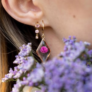 Könnte beinhalten: Goldfarbene Ohrringe mit rosa Edelsteinen und einem transparenten, rautenförmigen Anhänger mit einer kleinen, rosa Blume. Die Ohrringe werden in der Nähe eines lila Blumenstraußes gezeigt.