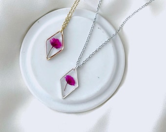 May include: Two necklaces with diamond-shaped pendants, each containing a pink flower. One necklace has a gold chain, the other a silver chain. The pendants are displayed on a white, circular dish against a white background.