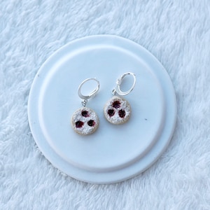 Könnte beinhalten: Ein Paar silberne Creolen mit einem kleinen, runden, weißen Keks-Anhänger mit roter Füllung. Die Keks-Anhänger haben einen weißen Puderzucker-Belag.