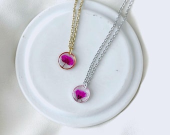 May include: Two delicate necklaces with round pendants, each featuring a vibrant pink flower encased in clear resin. One necklace has a gold chain, the other a silver chain. The pendants are displayed on a white, circular dish.