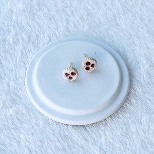 Puede incluir: Un par de pequeños pendientes redondos con fondo blanco y tres puntos rojos. Los pendientes están sobre una superficie blanca.