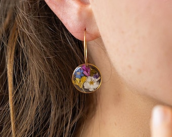 May include: Gold hoop earrings with a clear, circular pendant. The pendant is filled with a colorful assortment of dried flowers, including white, purple, blue, and yellow blooms. The earrings are shown on a person's ear.