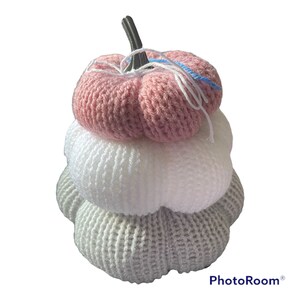 May include: A decorative stack of three knitted pumpkins in pink, white, and grey. The top pumpkin is pink, the middle one is white, and the bottom one is grey. A dark stem and white and blue yarn accents the top pumpkin. The pumpkins are stacked on top of each other.