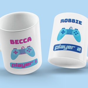 May include: Two white ceramic mugs with blue and pink gamer controller designs. One mug says "Becca Player 1" and the other says "Robbie Player 2".