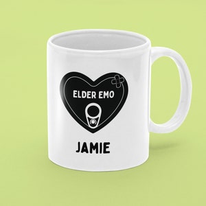 Pode incluir: Caneca de cerâmica branca com um design de coração preto que diz "Elder Emo" e um nome "Jamie" impresso abaixo do coração.