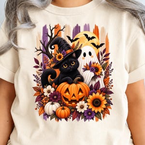 Peut inclure: Un t-shirt couleur crème arborant une illustration d'un chat noir portant un chapeau de sorcière, un fantôme blanc et une citrouille d'Halloween. Le motif est entouré de tournesols, de feuilles d'automne, de branches et de chauves-souris.