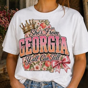 Peut inclure: T-shirt blanc avec un motif graphique. Le motif comprend le texte "She's From Georgia Y'all Can Tell", une couronne, des pêches, des éléments floraux et un nœud rose. Le t-shirt est en matière souple.