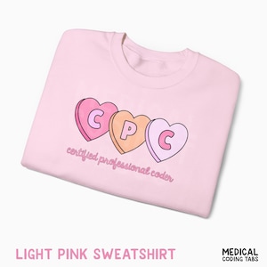 Peut inclure: Sweat-shirt rose clair avec un motif de trois bonbons en forme de cœur épelant "CPC" et les mots "certified professional coder" en cursive. Le sweat-shirt est de couleur rose clair.