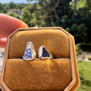 Puede incluir: Un anillo de plata con dos piezas distintas de vidrio marino engastadas en metal. Una pieza es un fragmento triangular de vidrio blanco con patrones azules, y la otra es una pieza triangular de vidrio azul oscuro sólido.