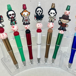 May include: A collection of pens with horror movie character toppers. Each pen features a different character, including Jason Voorhees, Pennywise, and Freddy Krueger. The pens have various colored barrels, including gold, green, and blue.