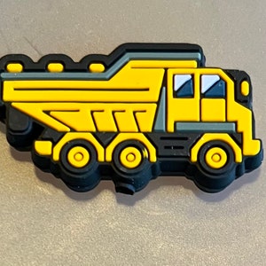 May include: A yellow and black construction truck charm. The charm features a detailed design of a dump truck with black wheels, a yellow cab, and a black outline. The truck has a gray top and two blue windows.