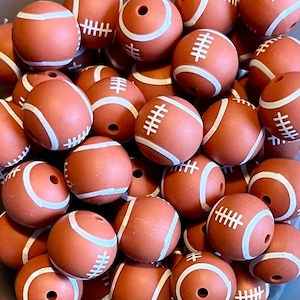May include: A close-up of a pile of brown silicone football-shaped beads with white stitching. The beads are perfect for making necklaces, bracelets, or other crafts.
