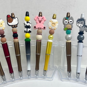 May include: A collection of ten decorative pens with animal-themed toppers. Each pen features a different farm animal, including a cow, sheep, chicken, pig, owl, and horse. The pens have a metallic finish and are displayed in a clear plastic holder.