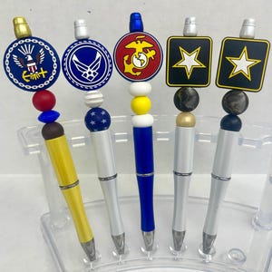 May include: Five pens with decorative tops featuring military emblems. The pens have various colors, including yellow, white, and blue, with silver accents. Each pen has a different design, including the Navy, Air Force, Marine Corps, and Army.