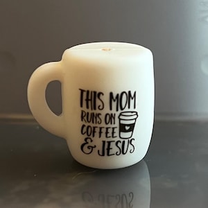 May include: A small, white mug-shaped charm with a handle. The mug has black text that reads "THIS MOM RUNS ON COFFEE & JESUS" and a graphic of a coffee cup. The charm is likely for a bracelet or necklace.