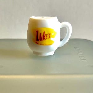 May include: White ceramic mug with a yellow and brown logo that says "Luke's" on it.