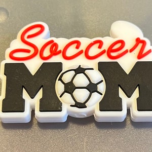 May include: A white and black Soccer Mom charm with a soccer ball in the center. The word "Soccer" is in red script above the word "MOM" in bold black letters. The charm has a white border.