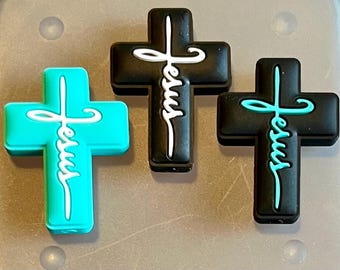 May include: Three cross-shaped charms with the word "Jesus" written in a stylized font. One charm is turquoise, one is black with white lettering, and the other is black with turquoise lettering.