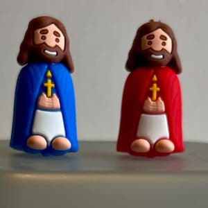 May include: Two small figurines of a person with a beard and long hair wearing a robe. One figurine is wearing a blue robe and the other is wearing a red robe. Both figurines are holding a cross in their hands.