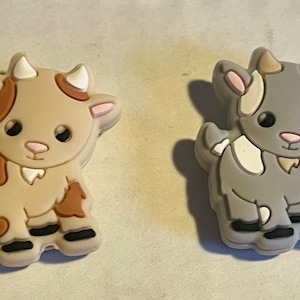 May include: Two cartoon goat charms. One is tan with brown spots, the other is gray with white spots. Both have black eyes, pink ears, and white horns. These charms are likely for shoes or other accessories.