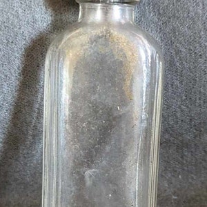 May include: A clear glass bottle with a black plastic lid. The bottle has a rectangular shape and is ribbed.