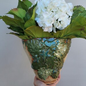 May include: A green ceramic vase with a textured floral design holds a bouquet of white hydrangeas and green leaves.