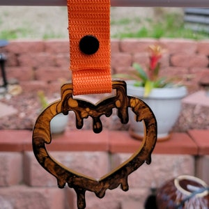 May include: A heart-shaped keychain with a dripping design in shades of brown and black, attached to an orange strap with a black button. The keychain is hanging in front of a blurred outdoor background.