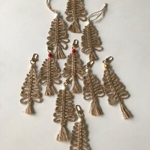 Könnte beinhalten: Eine Sammlung handgefertigter Weihnachtsbaum-Ornamente aus beigem Makramee. Jeder Baum hat ein geschlungenes Design, eine kleine Quaste am unteren Ende und einen goldfarbenen Verschluss oder eine Schnur zum Aufhängen. Einige haben rote oder goldene Perlen.