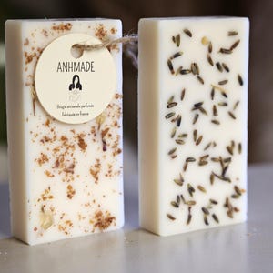 K&ouml;nnte beinhalten: Zwei rechteckige St&uuml;cke wei&szlig;e Seife mit unterschiedlichen botanischen Einschl&uuml;ssen. Die Seife links hat eine braune und wei&szlig;e gesprenkelte Optik mit einem kleinen Etikett, auf dem "ANHMADE" und "Bougie artisanale parfum&eacute;e fabriqu&eacute;e en France" steht. Die Seife rechts hat kleine, getrocknete Lavendelbl&uuml;ten, die in die Seife eingebettet sind.