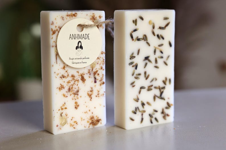K&ouml;nnte beinhalten: Zwei rechteckige St&uuml;cke wei&szlig;e Seife mit unterschiedlichen botanischen Einschl&uuml;ssen. Die Seife links hat eine braune und wei&szlig;e gesprenkelte Optik mit einem kleinen Etikett, auf dem "ANHMADE" und "Bougie artisanale parfum&eacute;e fabriqu&eacute;e en France" steht. Die Seife rechts hat kleine, getrocknete Lavendelbl&uuml;ten, die in die Seife eingebettet sind.