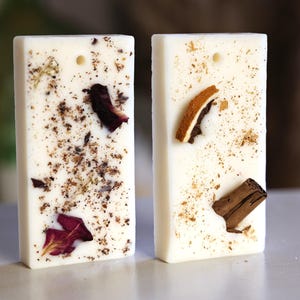 K&ouml;nnte beinhalten: Zwei wei&szlig;e Wachsschmelzen, eine mit getrockneten Blumen und die andere mit getrockneten Zitrusfr&uuml;chten und Zimt. Beide haben ein Loch zum Aufh&auml;ngen.