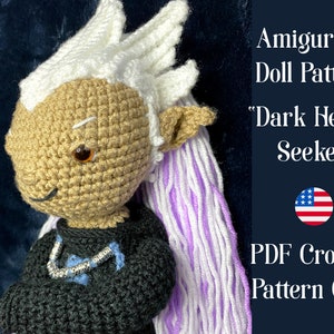 May include: Crochet amigurumi doll pattern for a character with white hair, a tan face, and a black shirt. The doll is wearing a silver necklace with blue beads. The pattern is called "Dark Hearts Seeker" and is available as a PDF download. The image also includes a US flag.