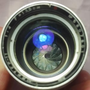 Può includere: Primo piano di un obiettivo fotografico con corpo argentato e nero. L'obiettivo presenta una serie di lamelle che formano un'apertura. Il vetro riflette luci blu e viola. L'obiettivo presenta le marcature "f=1:3.5 M" e "NP 6801".