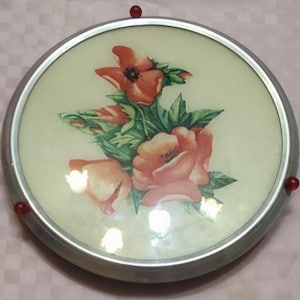 Puede incluir: Una bandeja redonda de color plateado con un diseño floral. La bandeja presenta un grupo de flores naranjas y rosas con hojas verdes. La bandeja tiene un borde elevado y cuatro acentos rojos alrededor del borde.