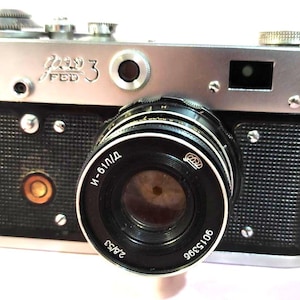 May include: A vintage FED 3 camera with a silver top and black body. The lens is black with white lettering, including "И-61Л/Д" and "2.8/53". The camera has various dials and buttons.