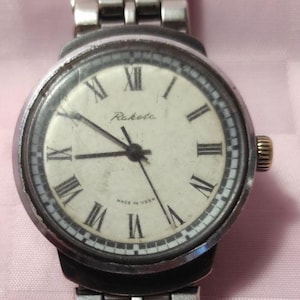 Puede incluir: Un reloj de pulsera Raketa vintage con una correa de metal plateado. La esfera del reloj es blanca con números romanos y agujas negras. Las palabras "Raketa" y "Made in USSR" son visibles. El cierre de la correa está marcado "CHINA".