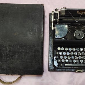 Puede incluir: Una máquina de escribir vintage negra con una funda de transporte negra. La máquina de escribir tiene un cuerpo de metal negro con una placa de identificación dorada que dice "Kappel".