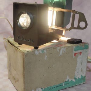 May include: A vintage slide projector in brown and green with a light bulb shining. The projector is sitting on a cardboard box with the text "ЭКРАН" in Cyrillic script.
