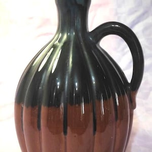 Puede incluir: Jarra de cerámica de dos tonos con tapa y asa. La parte superior es de color marrón oscuro, que pasa a una base marrón más clara. La tapa y el asa son de color marrón oscuro. La jarra tiene una forma bulbosa con estrías verticales.