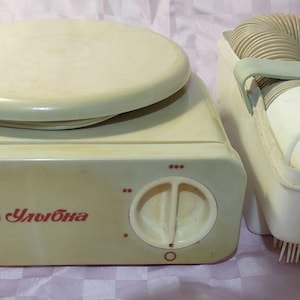 May include: An off-white, vintage foot warmer with a round top and a control dial. The unit is next to a matching foot drying device with a flexible hose and two foot-shaped inserts. The word "Улыбка" is printed on the front.