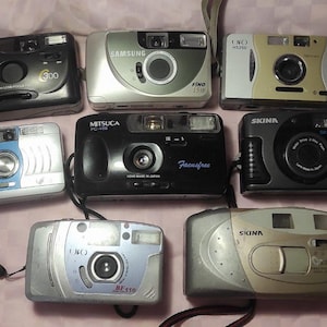 Puede incluir: Una colección de ocho cámaras de película vintage de apuntar y disparar en varios colores, incluyendo negro, plateado y dorado. Las marcas incluyen Samsung, Mitsuca y Skina. Algunas cámaras tienen correas de muñeca.