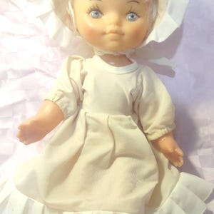 Könnte beinhalten: Eine Vintage-Puppe mit blondem Haar und blauen Augen, die ein cremefarbenes Kleid und eine Haube trägt. Das Kleid der Puppe hat einen Rüschensaum und die Haube ist gefaltet. Die Haut der Puppe hat einen hellpfirsichfarbenen Farbton.