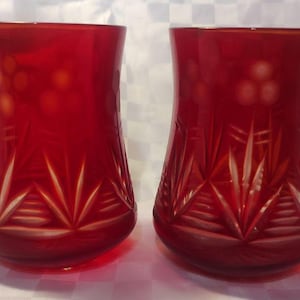 May include: Two ruby red, cut-glass tumblers. The glasses have a flared shape and feature intricate starburst and floral designs. The cut glass details catch the light, creating a sparkling effect. These decorative glasses are ideal for serving beverages.