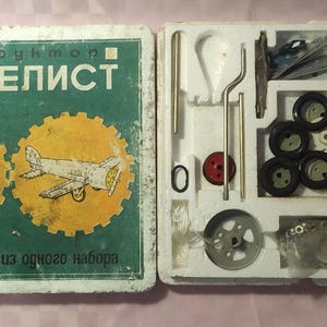 Puede incluir: Una caja de kit de maquetas vintage con el texto "Конструктор Моделист" en la parte delantera. La caja contiene una variedad de piezas metálicas, incluidas ruedas, ejes y engranajes. La caja también incluye instrucciones para construir un modelo.