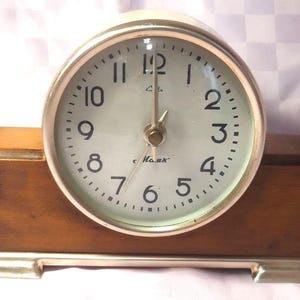 May include: A vintage desk clock with a round, cream-colored face and black numerals. The clock has a silver-toned bezel and hands, and is set in a rectangular wooden base with a silver-toned trim. The clock reads "c. Max" on the face.