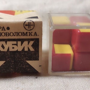 Può includere: Un cubo di puzzle in plastica rosso e giallo in una scatola di plastica trasparente. La scatola ha il testo "Игра - Головоломка" e "Кубик" stampato su di essa.