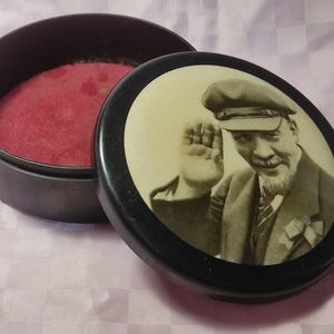 May include: A vintage round black box with a lid featuring a sepia-toned portrait of a man in a cap, waving. The box is open, revealing a red velvet interior. The lid has a black border.