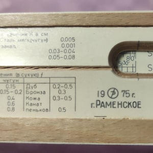 Może przedstawiać: Zbliżenie na zabytkową drewnianą suwakową linijkę z wydrukowanymi tabelami i skalami. Suwakowa linijka ma zakrzywione wycięcie i tekst, w tym "Sin" i "S&T". Widoczny jest również tekst "19 75 r. г. РАМЕНСКОЕ".