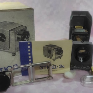 May include: A vintage slide projector with a silver and black design. The projector is in its original box with a manual and other accessories. The box is labeled "Этюд 2с".